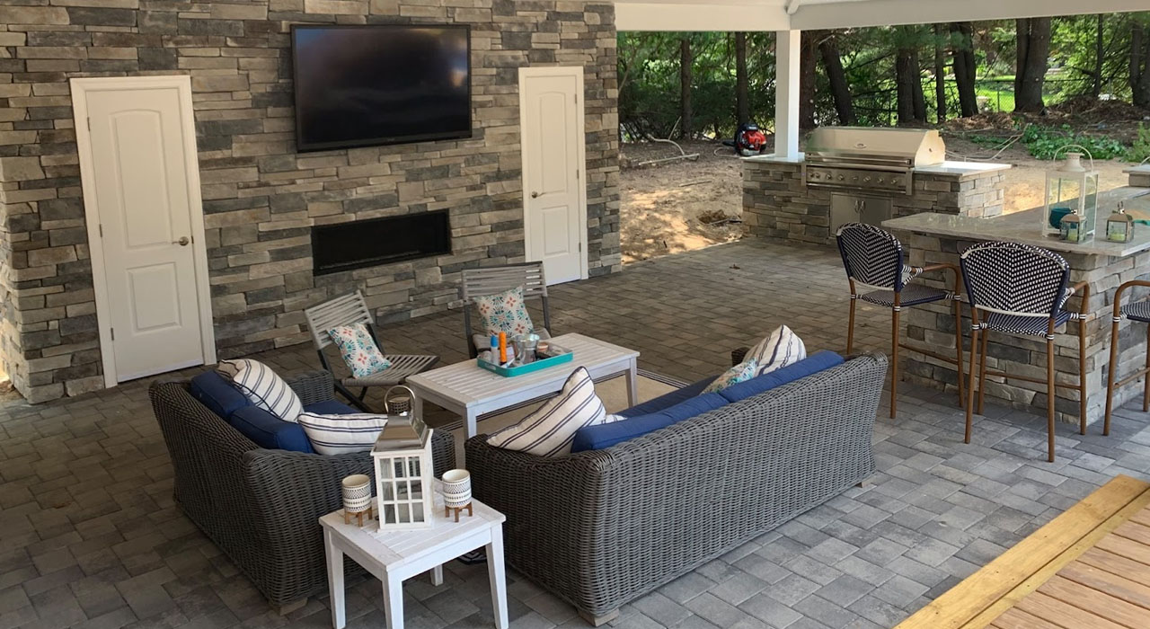 Covered outdoor living area featuring stone walls, a paver floor, and built-in kitchen, showcasing premium finishes that raise the patio cost per square foot.