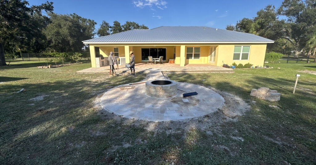 Backyard patio installation featuring circular fire pit, curved paver walkway, and crew working near porch.