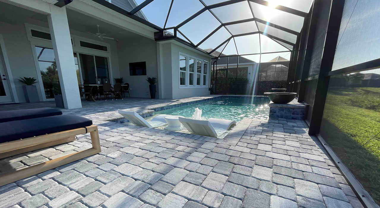 Modern pool patio with paver decking, in-water loungers, and custom lanai enclosure providing shade.