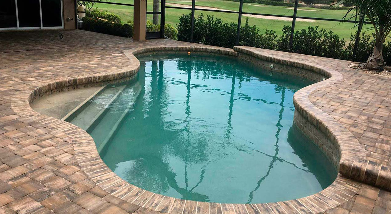 Curved swimming pool surrounded by brick pavers inside a screened lanai enclosure with clean finished edges.