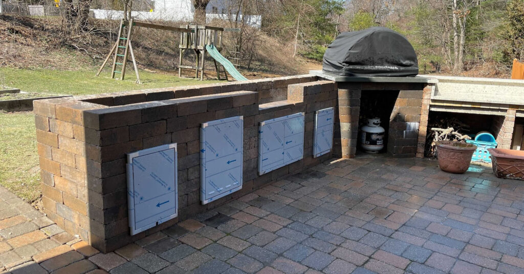 Stone outdoor kitchen with built-in oven, storage doors, and patio pavers in a backyard setting.