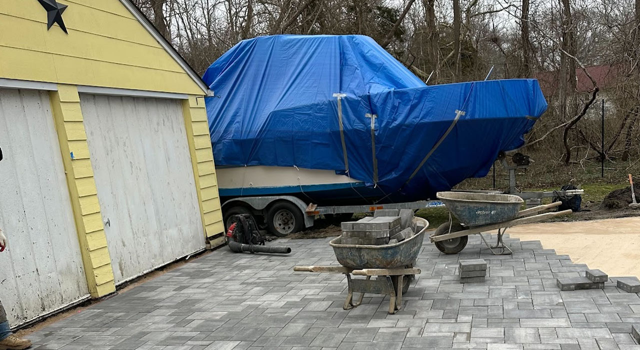 Paver driveway installation in progress beside garage, with wheelbarrows, stacked pavers, and covered boat nearby.