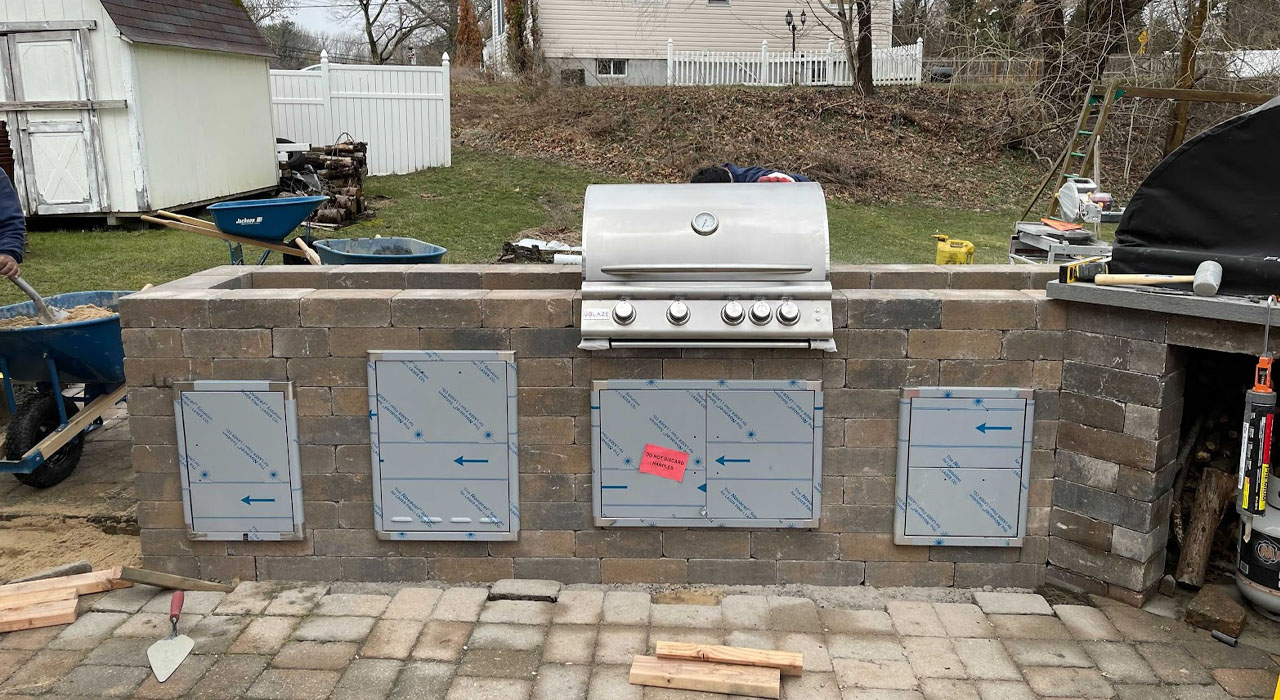 Completed outdoor kitchen section featuring stainless steel grill, storage cabinets, and finished stonework.