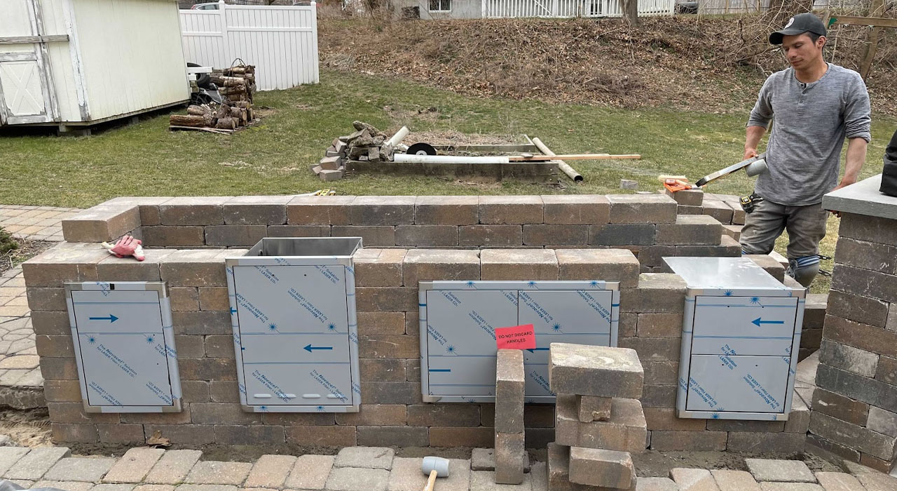 Masonry outdoor kitchen wall under construction with access panels and worker installing stone blocks.