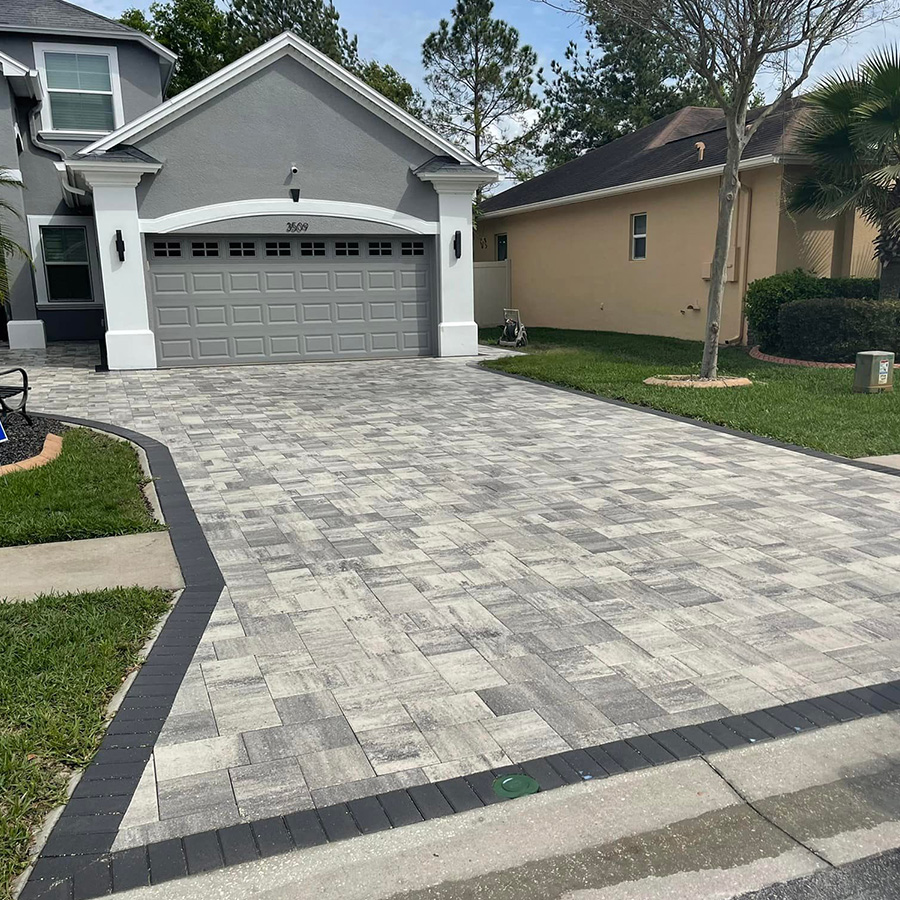 This gray stone driveway features multi-toned pavers arranged in a textured, random pattern. A dark charcoal border outlines the path, which leads to a gray garage with white trim and pillars.