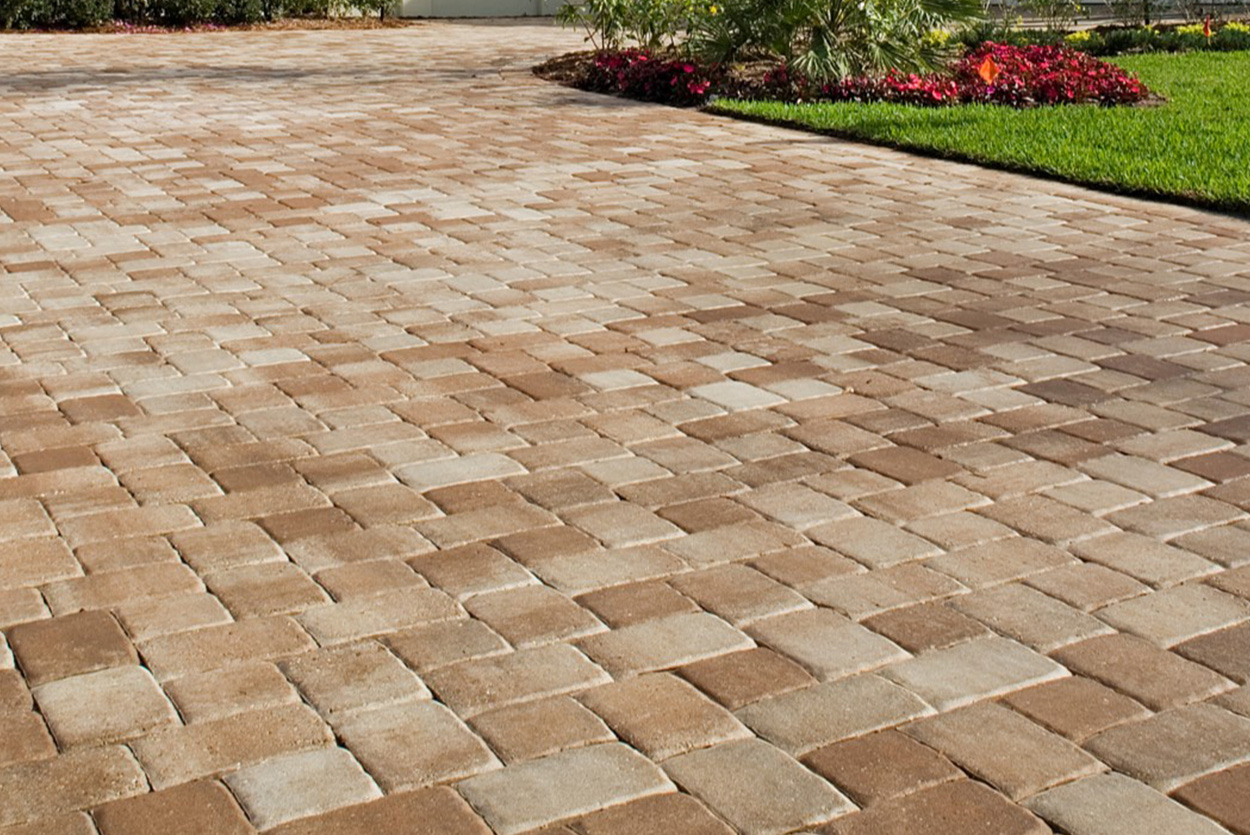 This sprawling driveway is composed of small, rectangular stone pavers in warm shades of tan and light brown. The stones are arranged in a neat running bond pattern, leading past vibrant red flowers.