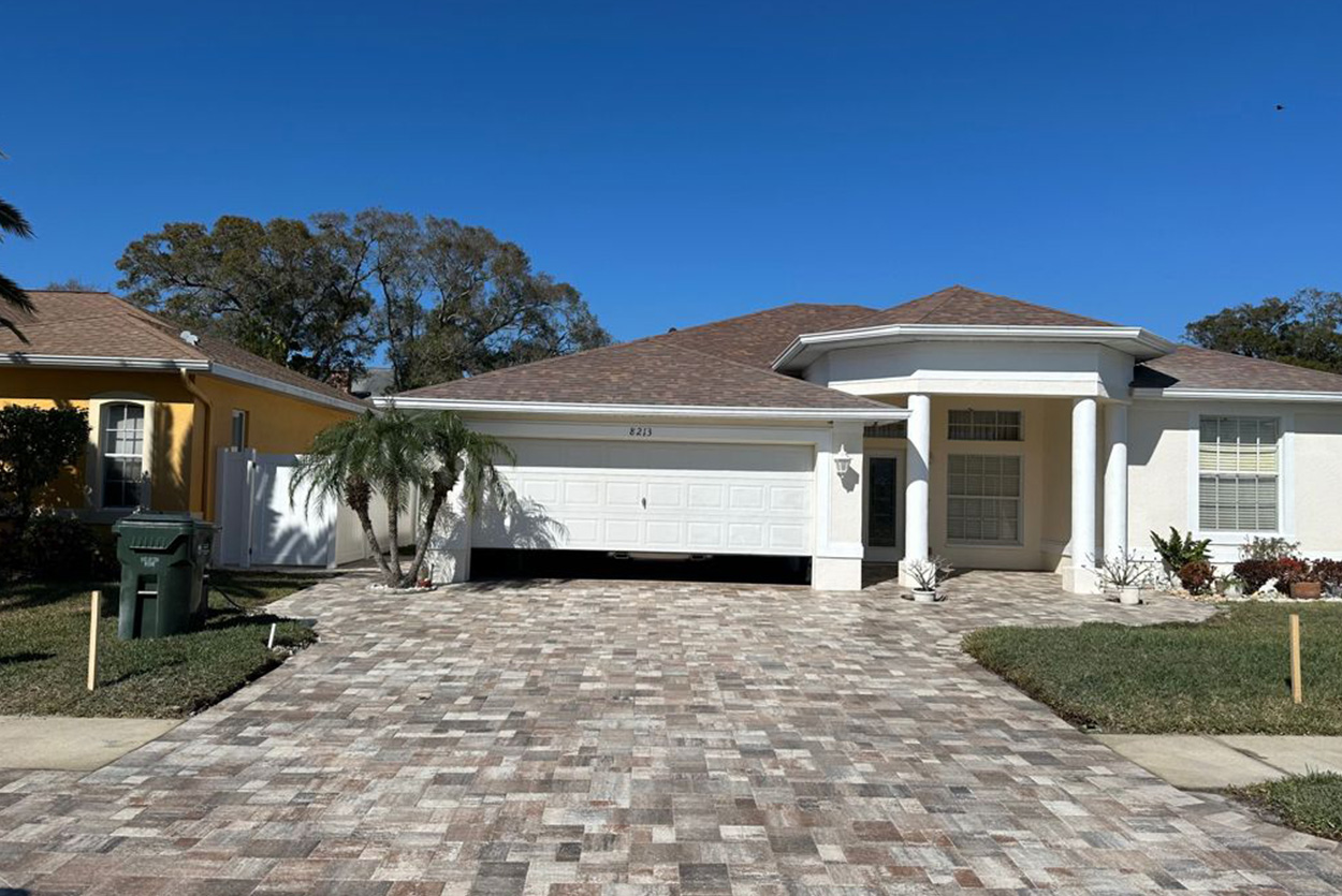 This single-story white house features a large driveway and entryway paved with multi-toned brown and gray stone pavers. A white garage door and large white pillars frame the entrance under a clear blue sky.