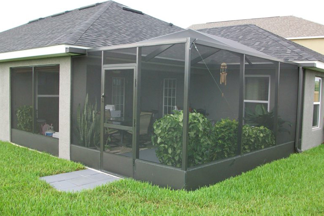 This screened-in lanai features a dark metal frame and a pitched roof, enclosing a patio with several leafy green plants inside. A small gray stone walkway leads from the lush green lawn to the screen door.