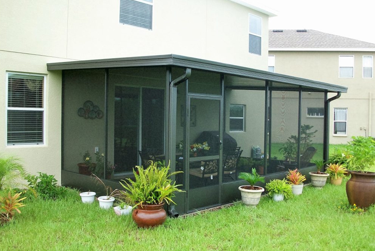 This screened-in lanai features a roof extension and a dark metal frame with mesh walls. Various potted plants, including ferns and colorful bromeliads, line the exterior base of the structure.