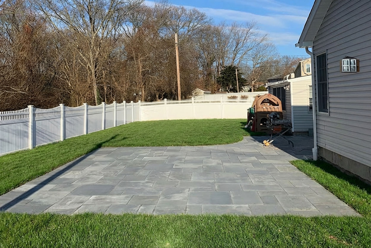 This unique hardscape design features a large rectangular patio section seamlessly transitioning into a graceful curved edge. Uniform grey pavers extend across the manicured lawn, bordered by a decorative white vinyl fence.