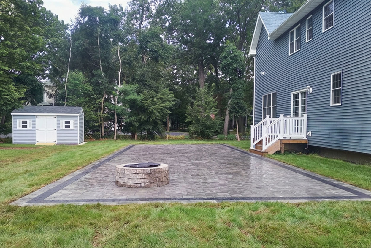 Expansive backyard patio features multi-toned grey pavers highlighted by a bold black border. A round, stacked-stone fire pit serves as a central focal point within the professionally landscaped lawn.