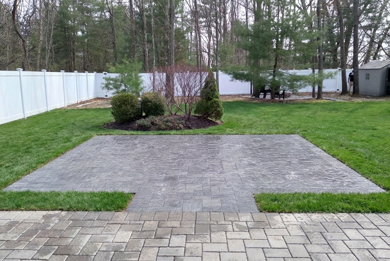 Backyard landscape features two connected square patios showcasing distinct paving styles. A light grey rectangular paver area transitions into a darker, textured stone section centered within the manicured lawn.