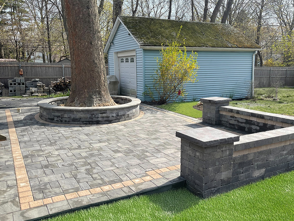 Professional hardscape features a mature tree centered within a circular, stacked-stone retaining wall. The surrounding multi-toned grey patio is accented by a tan border and matching L-shaped sitting walls.