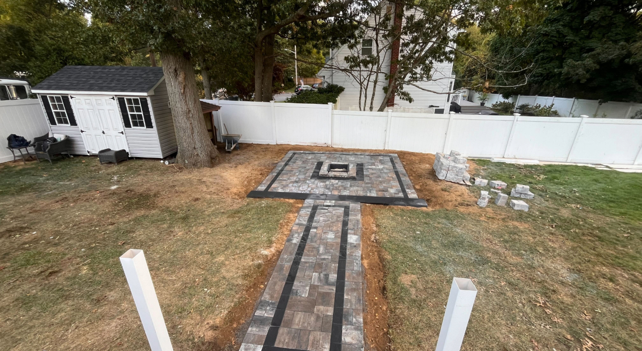 A square stone fire pit built into a matching paver patio with a dark border, demonstrating a custom fire pit installation.