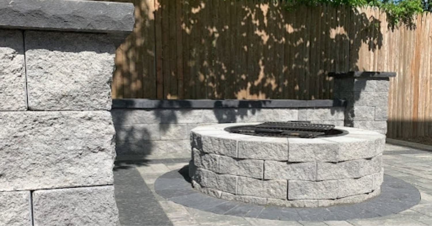 A circular grey stone fire pit with a grill grate and matching seating wall, showing a completed fire pit installation.