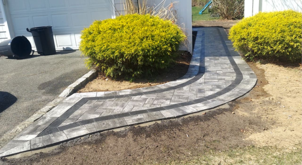 A curved stone path with a dark border wrapping around front yard bushes, demonstrating a custom walkway installation.