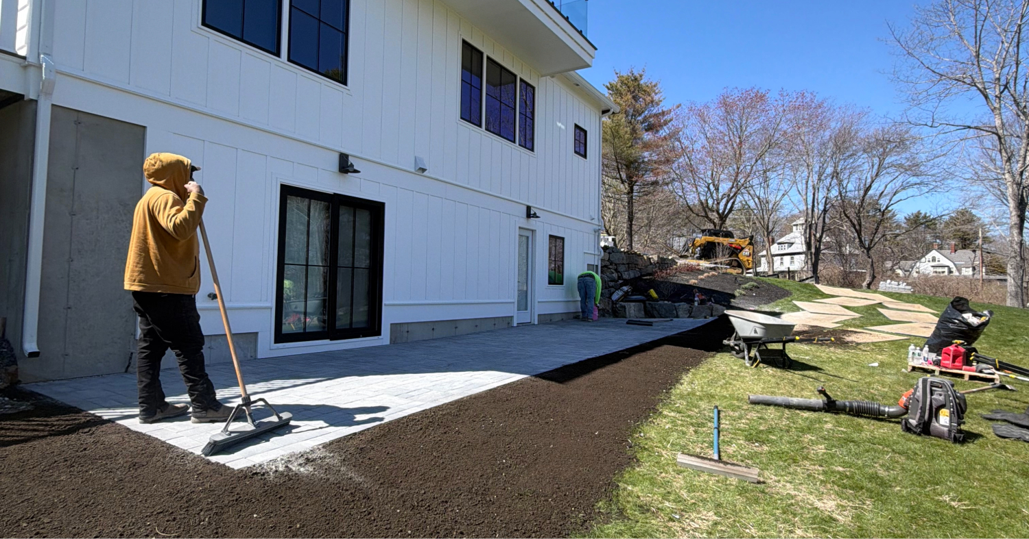 A landscaper carefully levels rich topsoil next to newly installed pavers, showcasing creative patio ideas in Manchester for large backyards.