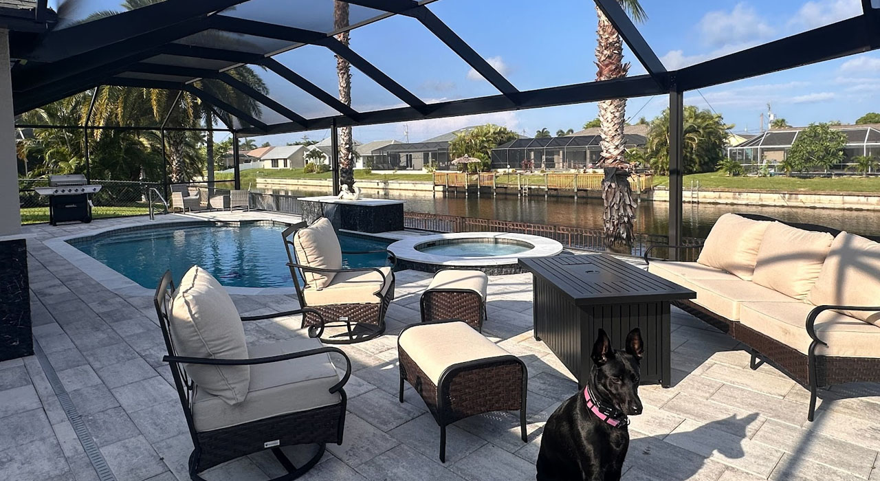A cute black dog sits on a paved patio, enjoying the water view inside one of many pool screen enclosures.